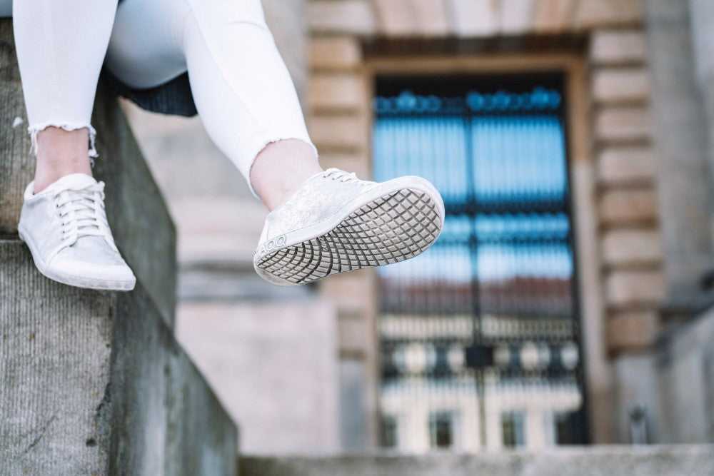 Eine Person trägt die stylischen TIQQ Silver White Sneaker von ZAQQ aus hochwertigem Nappaleder, kombiniert sie mit weißen Hosen und sitzt mit baumelnden Beinen auf einem Felsvorsprung. Im Hintergrund ist eine verschwommene Ansicht eines Gebäudes mit großen Eisentoren zu sehen.