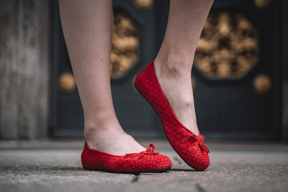 Eine Person, die ZAQQs TWIST Red Ballerinas mit gewebtem Design trägt, steht auf einem Steinpflaster. Im Hintergrund ist eine dunkle Tür mit aufwendigen goldenen Mustern zu sehen, die die Eleganz des Velourleder-Finishs unterstreichen.