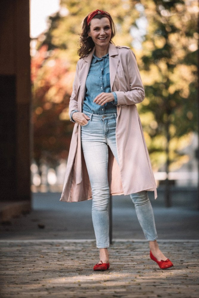 Eine Frau steht draußen auf einem Kopfsteinpflasterweg. Sie trägt einen hellrosa Trenchcoat, ein Jeanshemd und Jeans. Dazu trägt sie ein rotes Stirnband und schicke Ballerinas aus der TWIST Velours Red-Kollektion von ZAQQ und lächelt vor einem verschwommenen Hintergrund aus Grün und Bäumen.