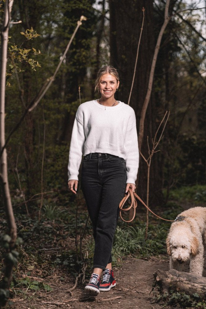 Eine Frau in weißem Pullover und schwarzer Hose führt einen flauschigen Hund an der Leine durch einen Wald. Ihre Füße stecken fest in roten, wasserdichten Schuhen von ZAQQ VAQATION. Sie lächelt, umgeben von Bäumen und üppigem Grün.