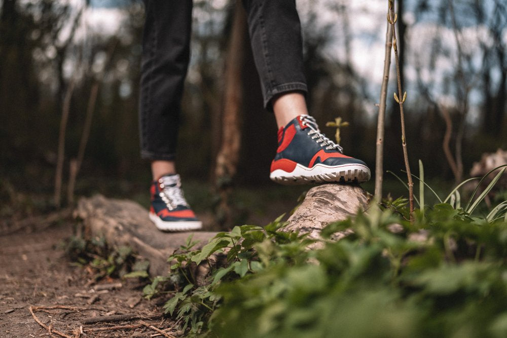 Eine Person in schwarzer Hose und ZAQQs VAQATION Red Waterproof Sneakers mit der strapazierfähigen TERRA-Sohle läuft auf einem umgestürzten Baumstamm in einem Wald. Der Weg ist von Grün umgeben und der Hintergrund ist unscharf, was eine ruhige, natürliche Atmosphäre schafft.