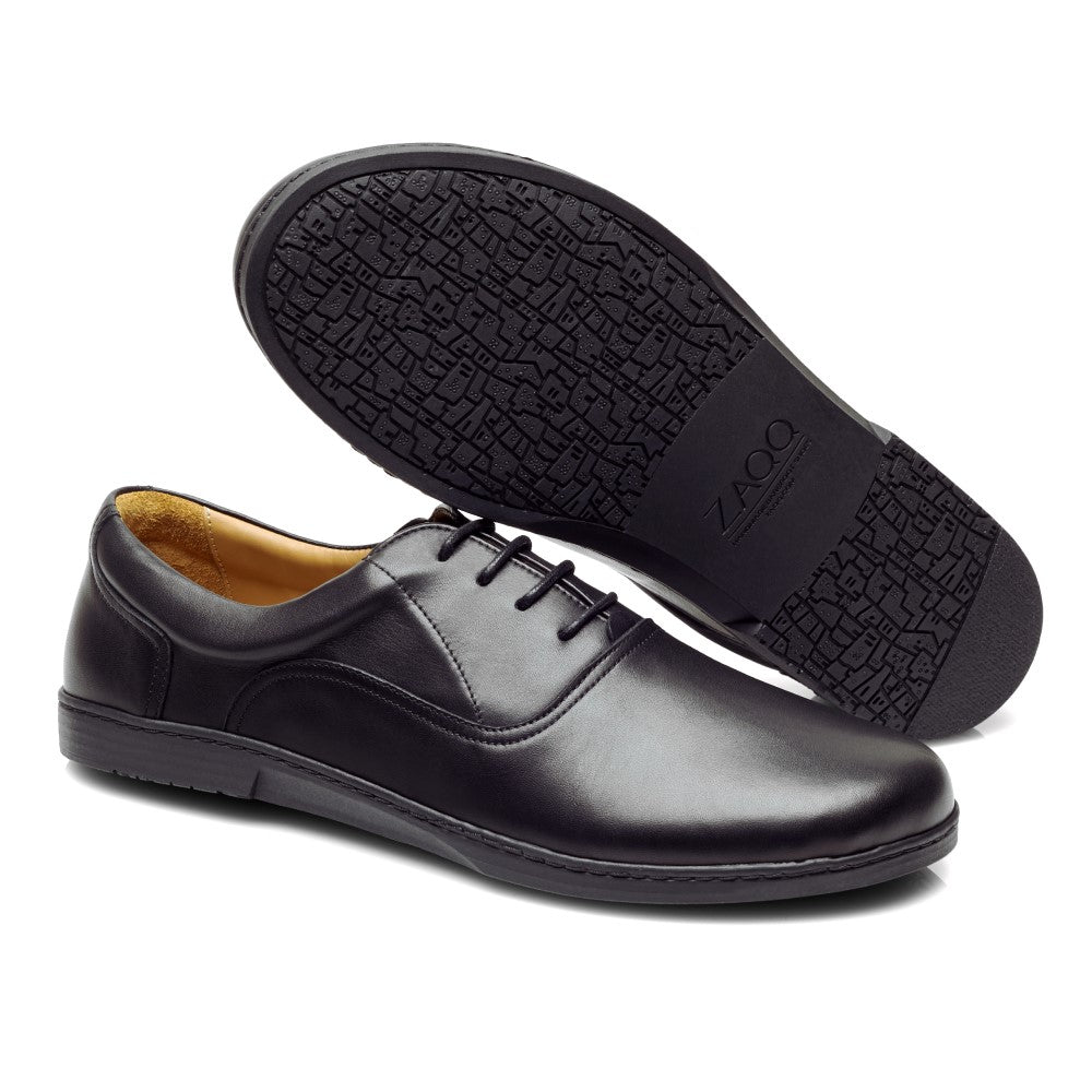 Ein Paar elegante APEQ Black-Schuhe von ZAQQ, gefertigt aus feinem Kalbleder. Ein Schuh liegt flach mit sichtbaren Schnürsenkeln und Nähten, während der andere geneigt ist, um die strukturierte Sohle und das Markenlogo zur Geltung zu bringen.