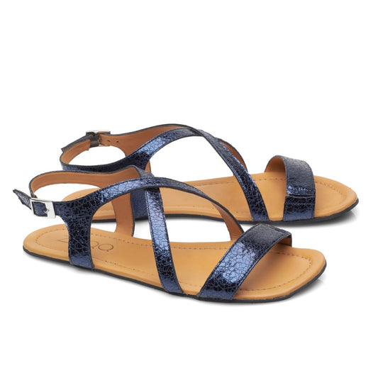 Ein Paar ZAQQs QEE-Blue Barfuß-Sandalen für Damen mit überkreuzten Riemen in dunklem Metallic-Blau über den Zehen und einer hellbraunen Sohle, abgerundet durch einen Knöchelriemen und Schnallenverschluss, alles aus weichem Nappaleder gefertigt.